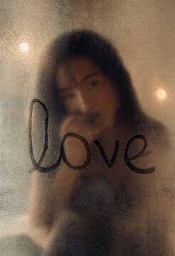 Emotional woman looking through steamy window with the word love written on it