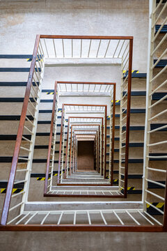 Vertical top-down view of a multistory square stairwell with repeating stairs and railings, creating strong geometric perspective, symmetry, depth and an abstract architectural pattern.