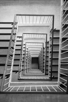 Black and white top-down view of a multistory square stairwell with repeating stairs and railings, creating strong geometric perspective, symmetry, depth and an abstract architectural pattern.