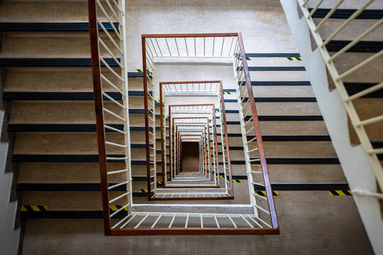 Top-down view of a multistory square stairwell with repeating stairs and railings forming a geometric spiral, strong perspective and deep architectural pattern inside a building.
