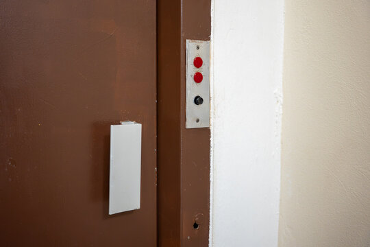 Close-up of an old elevator door with handle and vintage call button panel, showing red and black controls, worn equipment and aging vertical transportation infrastructure.