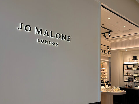 A sleek interior view of a Jo Malone store in London. The shop features a minimalist design with a focus on white walls and soft lighting. Display shelves are lined with luxury fragrance products.