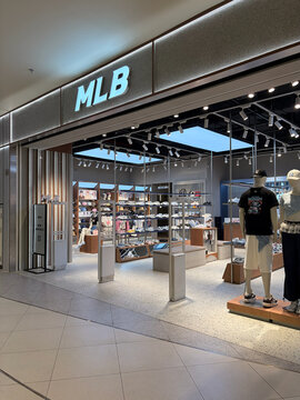 A retail store entrance with the MLB logo prominently displayed. The glass storefront showcases sports apparel and merchandise. The interior is well-lit, featuring shelves and mannequins.