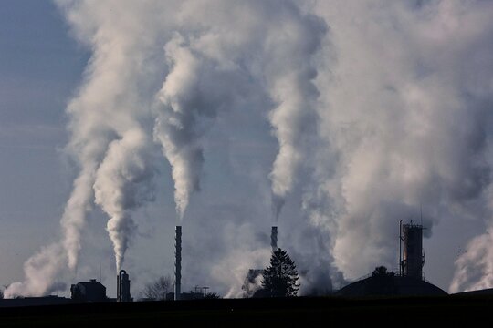 Vue de loin d'une usine avec de hautes chemin&eacute;es align&eacute;es desquelles jaillit une &eacute;paisse fum&eacute;e blanche qui se disperse en hauteur sous forme de colonnes dans le ciel gris&acirc;tre. Symbole de la pollution.
