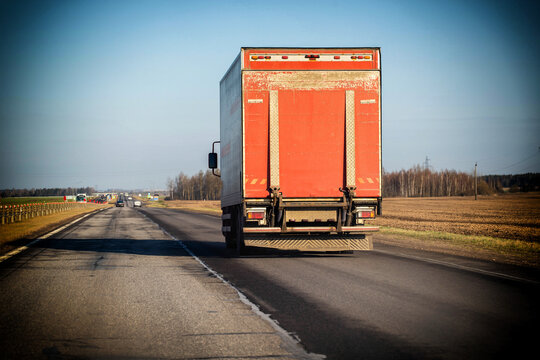 Red cargo truck trailer on open highway driving away, transport vehicle movement on road, clear blue sky background, rural landscape of cultivated fields.