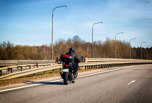 Motorcycle journey on open highway under clear sky. Rear view of rider on black motorbike with top case, passing roadside barrier and leafless trees.