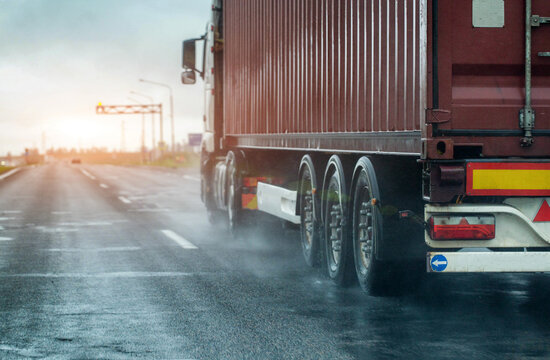 Heavy cargo truck with shipping container travels on wet highway, rear view of wheels splashing water, indication of slippery road condition and reduced visibility due to rain. Container truck