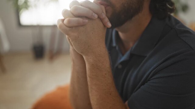 Man, young hispanic with beard and curly hair, hands clasped and pressed to lips in prayer on a bed in a studio; pensive solitude.