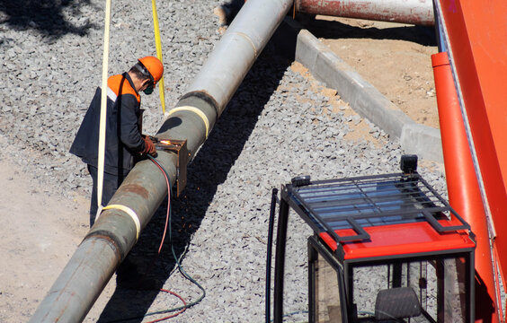 Construction worker welding metal bracket to large industrial pipeline at outdoor site under bright sunlight. Safety gear worn by worker during pipe installation process.
