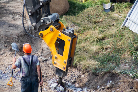 Construction worker with shovel observes hydraulic hammer excavator attachment breaking concrete on outdoor site, showing heavy machinery power and labor work process. Copy space