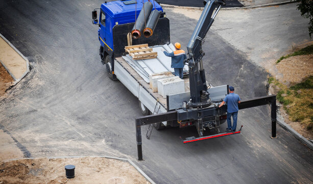 Blue construction truck equipped with hydraulic manipulator crane system unloads large insulated pipes for infrastructure project. Two workers assist with heavy material handling on road construction