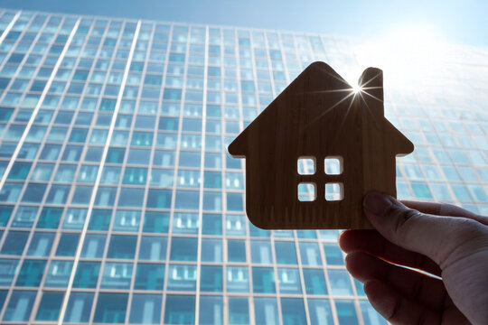 Real estate and modern architecture concept with hand holding wooden house model against glass skyscraper condominium building with sunburst for city apartment buying and property investment.