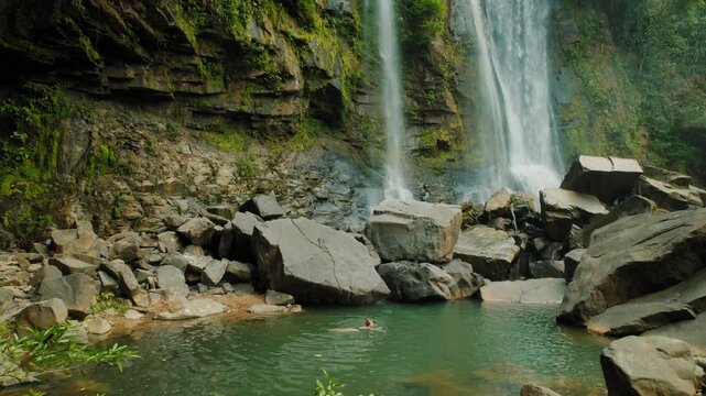 Traveler swimming in clear pool beneath cascading waterfall and rocky cliffs