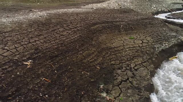 A critically low, muddy stream of water struggles to flow through a vast, severely cracked and dry riverbed, shrinking natural resources, severe drought conditions