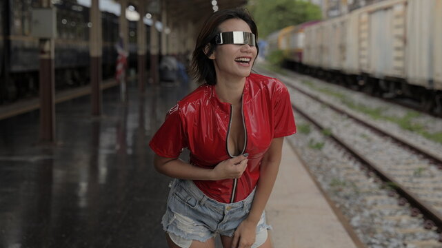 A playful Asian female model wearing metallic visor eyewear, an unzipped red vinyl top, and denim shorts poses with a wide smile on a train station platform