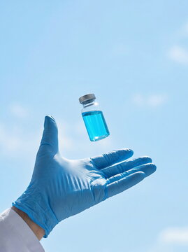 Medical vial with blue liquid floating above gloved hand against bright sky, symbolizing scientific innovation.