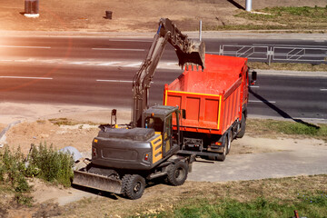 Excavator loading soil into red dump truck at construction site near highway. Heavy machinery moving earthwork material for road infrastructure development project, copy space © HENADZY