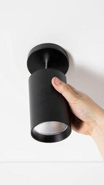 Hand installing modern black cylindrical LED spotlight fixture on white ceiling. Electrical work, home improvement, lighting installation, interior design project vertical photo
