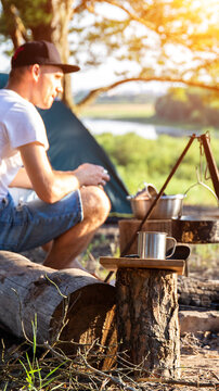 Adult male camper crouches near campfire setup with cooking pots and metal mug outdoors at sunny campsite by river, tent and lush foliage in background, vertical photo