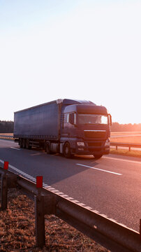 Dark blue semi truck with cargo trailer driving on highway during sunrise, golden light on road surface and guardrail, clear sky, vehicle transporting goods across country vertical photo