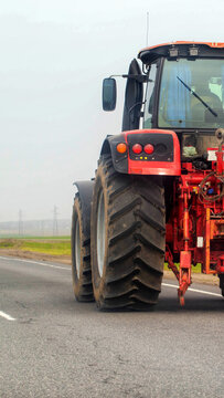 Red agricultural tractor driving on asphalt road, large muddy tires, rear and side view of heavy vehicle, rural landscape with fields and power lines background, overcast sky vertical photo