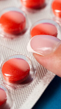 Human finger pressing bright red pill from aluminum foil and plastic blister pack, close-up view of medicine preparation, health care and pharmaceutical concept, vertical photo