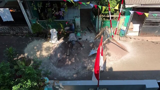 Time Lapse video of Man stirring sand in sunlit courtyard. Red flags flutter beside him gently. Children watch from doorway, smiling. Shadows dance on dusty floors. Scene evokes warmth and tradition. 
