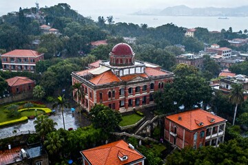 Obraz premium Aerial view of a historic red-brick building with a dome, surrounded by greenery and residential structures near a coastal area during rainfall