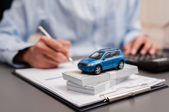 Auto loan car insurance and vehicle finance purchase concept. Blue miniature toy car on stack of dollar money cash with man calculating on document.