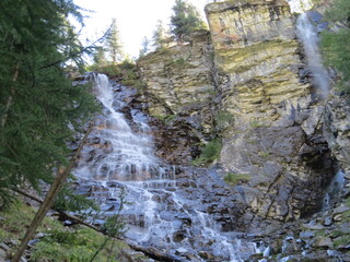 A scenic alpine waterfall cascades down rugged rocky cliffs surrounded by evergreen pine trees in summer. © Roberto