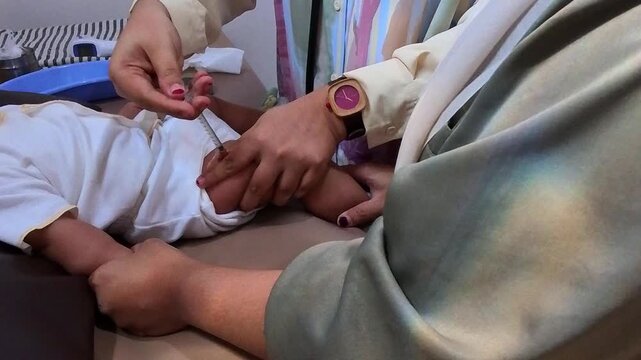 Video of Healthcare worker administering an injection to a baby. The child lies on a bed, calm and still. The worker's hands gently hold the baby's legs. Watch adorns the worker's wrist. Care and prof