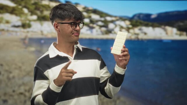 Man pointing index finger at a white chocolate bar while holding it up near chest in studio; playful curiosity.