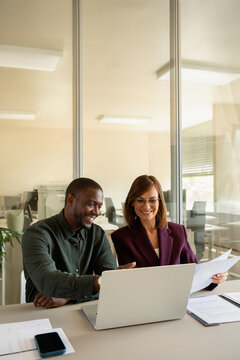 Vertical photo of business man and woman working on laptop in office, holding paper document. Diverse multiracial colleagues analysing company report online together, copy space, team in 30s and 40s.