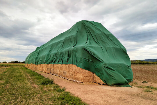 Huge multi‑level stack of square hay bales partly covered with a green tarp under a grey sky