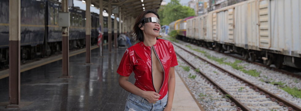 Playful Asian woman wearing futuristic metallic visor sunglasses and a red unzipped top stands at a railway station platform