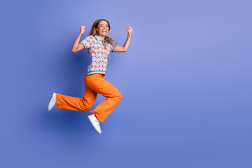 Fototapeta premium Happy young woman leaps in mid air against a blue background wearing orange pants and a patterned polo top