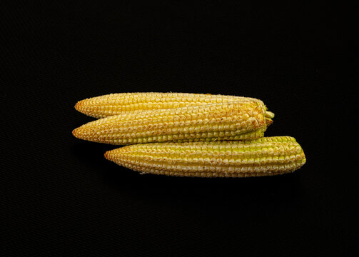 Three mini corncobs with yellow kernels arranged diagonally on black textured background, shallow depth of field, studio lighting