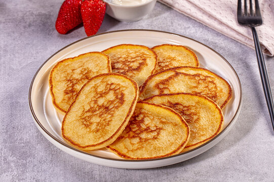 Golden mini pancakes on white plate with fresh strawberries and cream, rustic table setting, soft natural lighting