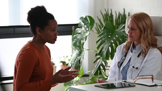 Concerned Black woman describing symptoms to female physician during medical appointment. Doctor listens carefully during professional healthcare consultation