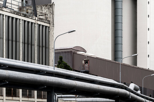 Industrial pipes and factory architecture form layered urban infrastructure beneath an overcast skyline with modern buildings in the background