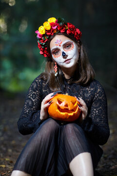 Woman with Santa Muerte make-up sits on a path in forest and holds a Halloween pumpkin in hands.