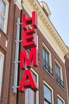 A prominent red HEMA logo is mounted vertically on the facade of a traditional brick building under a clear sky. The HEMA is a Dutch variety chain store known by low priced household goods.