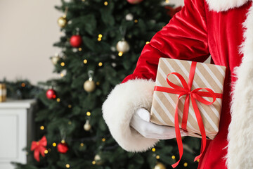 Obraz premium Christmas gift giving and holiday celebration, Santa Claus holding a wrapped present with a red ribbon in front of a decorated pine tree at home
