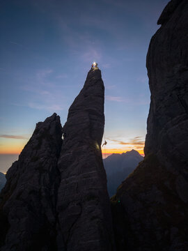 Aerial view of Punta Carina rock spire at twilight with a climber rappelling down, illuminated by headlamps against a colorful sunset sky in Minucciano, Toscana, Italy.
