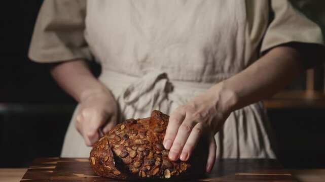 Close-up video of a baker wearing a rustic linen apron slicing fresh homemade artisan bread topped with pumpkin seeds on a dark wooden cutting board. Perfect for bakery promos, healthy food concepts.