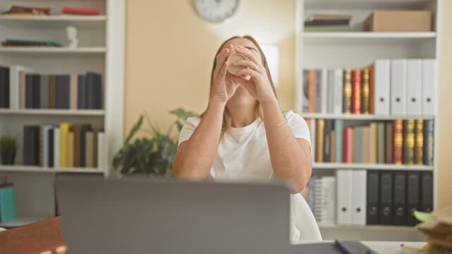 Woman at laptop tucks hair with hand and sips coffee while reading a book in a library building; concentration productivity.
