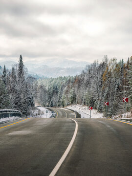 Beautiful empty highway road with road signs in winter pine forest. Snowy hills and mountains under cloudy sky. Winter asphalt road. Vertical scenic nature background with copy space. Seasonal travel