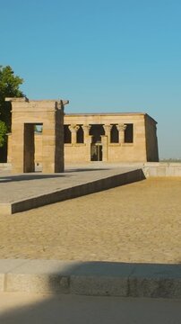 A low angle shot of the historic Egyptian Temple of Debod in Madrid, Spain, featuring ancient stone gateways and lush green trees under a clear blue sky.