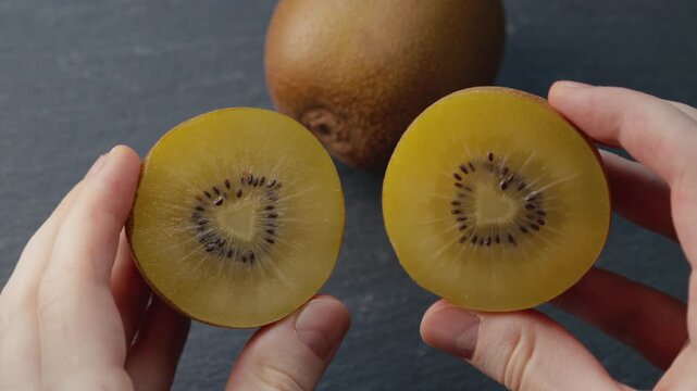 Macro shot of male hands holding two halves of a ripe golden kiwi and gently rotating them on gray background. Rich juicy pulp and detailed texture for healthy eating, gourmet food, culinary content.