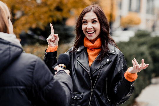 Smiling woman interviewed outdoors in autumn park wearing black leather jacket and orange turtleneck, gesturing and speaking to a reporter holding a microphone